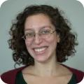 Smiling woman with curly hair and glasses wearing a red and green top against a dark background.
